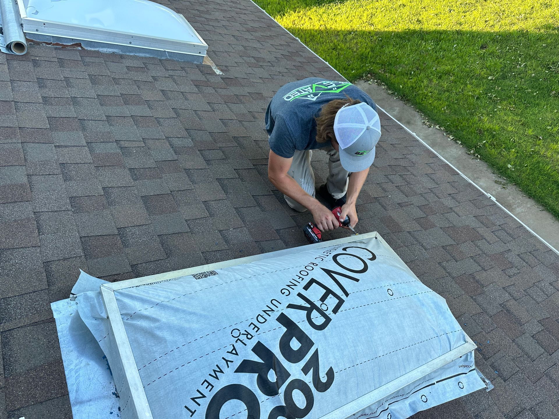 A person on a brown shingled roof cutting material with a power tool near a stack of underlayment.