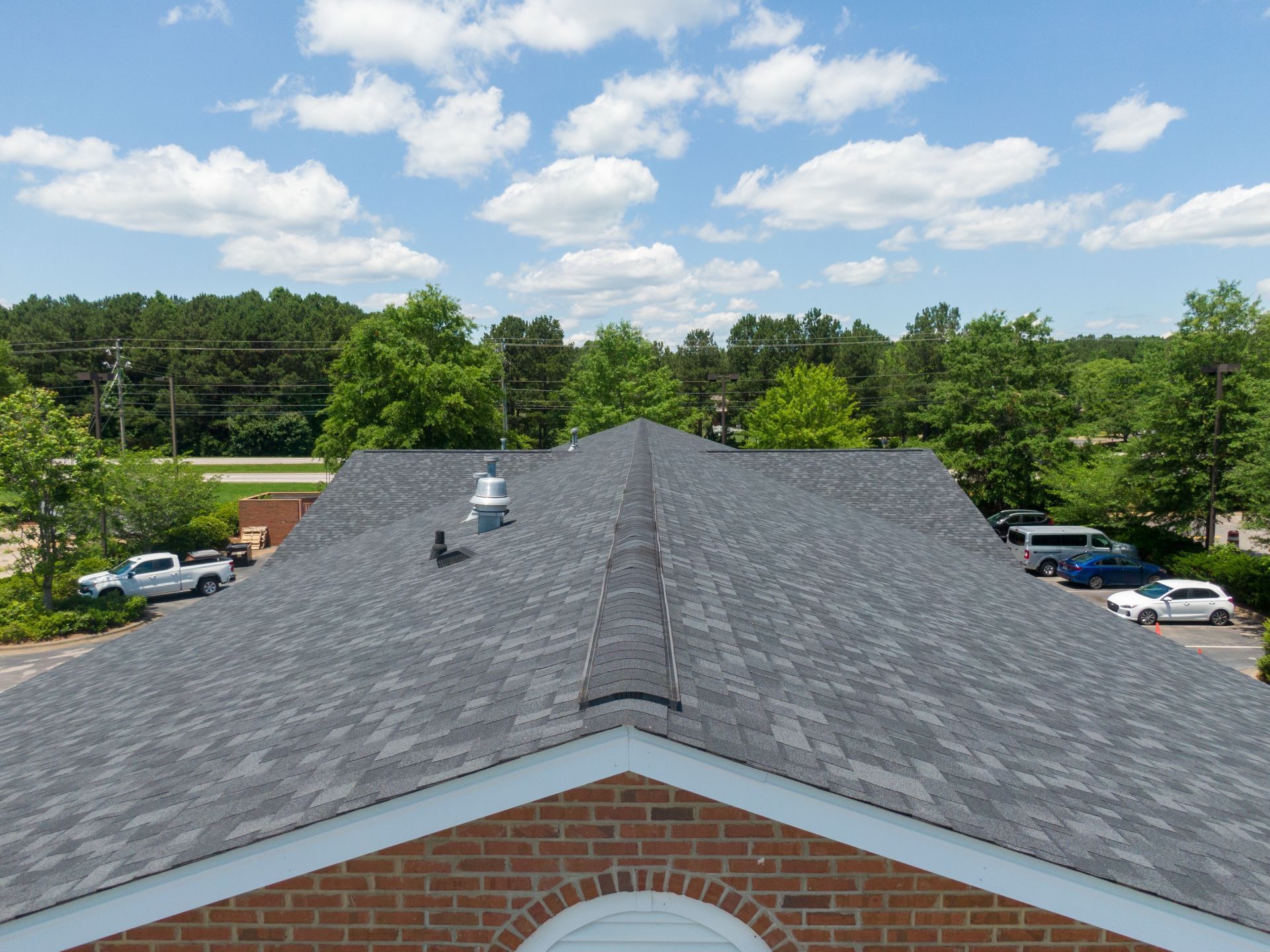 Gray shingle roof on a brick building, under a blue sky with clouds. Trees and vehicles visible in background.