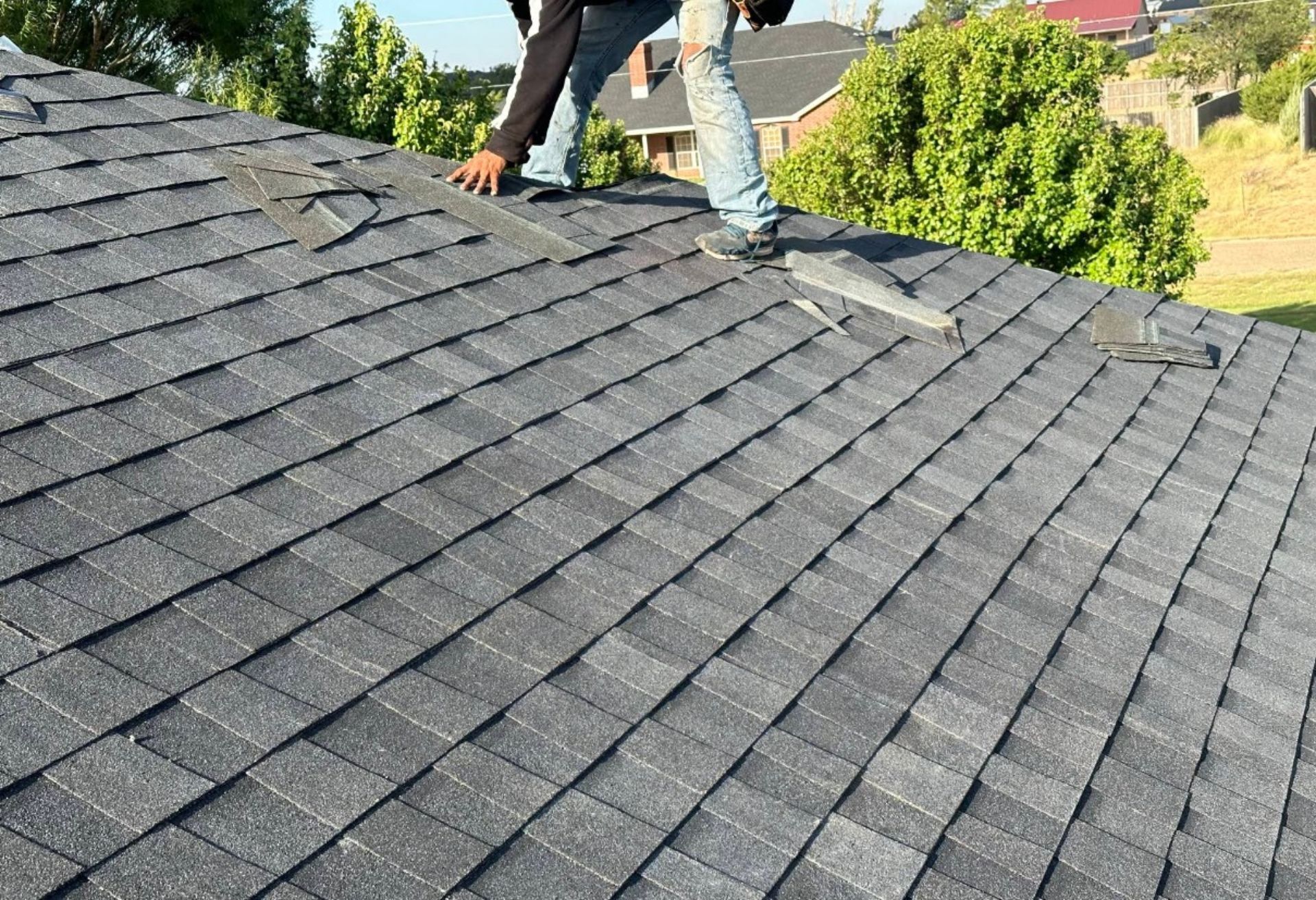 Person on a gray shingled roof inspecting damage. Blue jeans, green trees, sunny day.