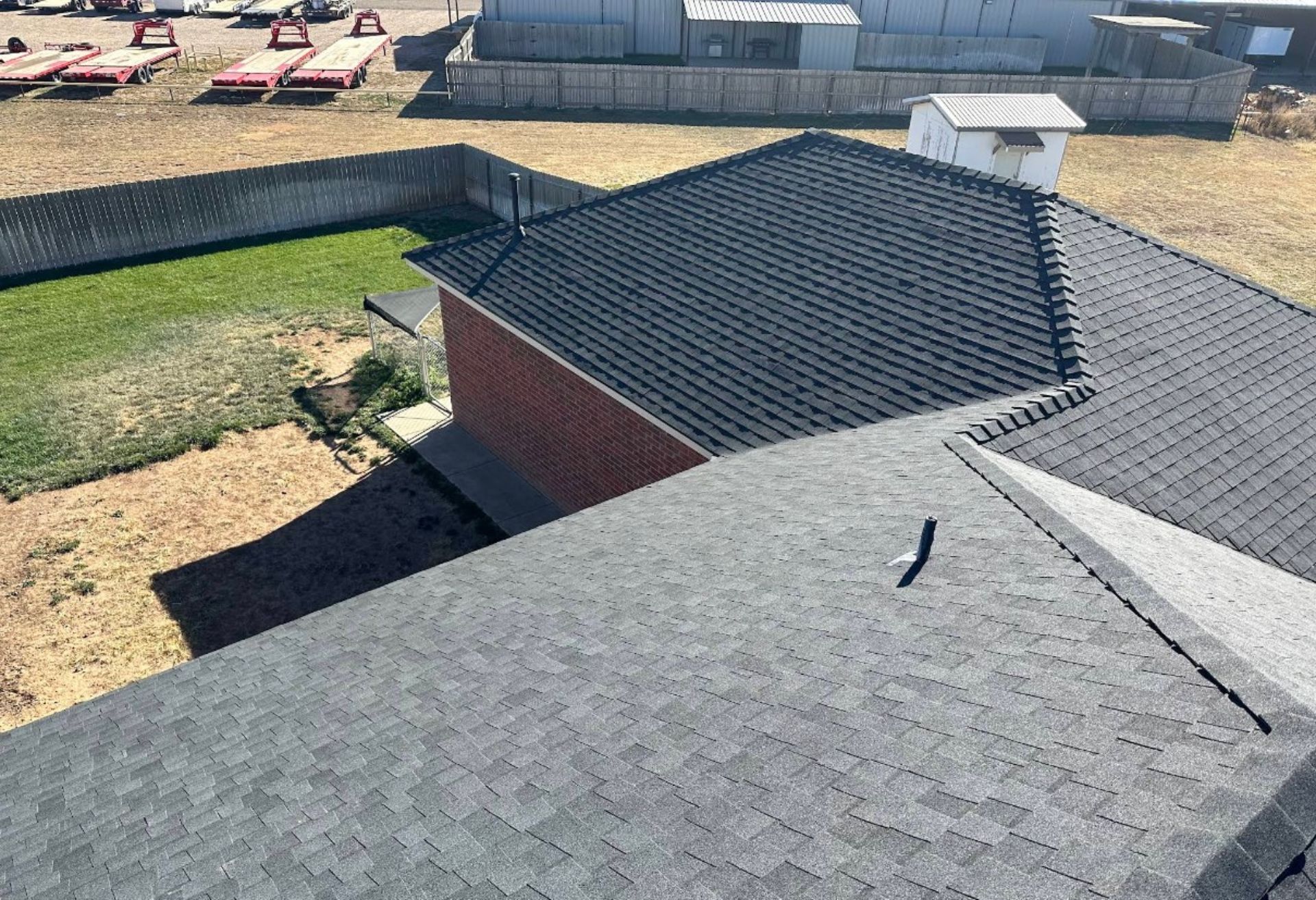 Aerial view of a dark shingled roof next to a brick wall and grassy area. The setting is sunny.