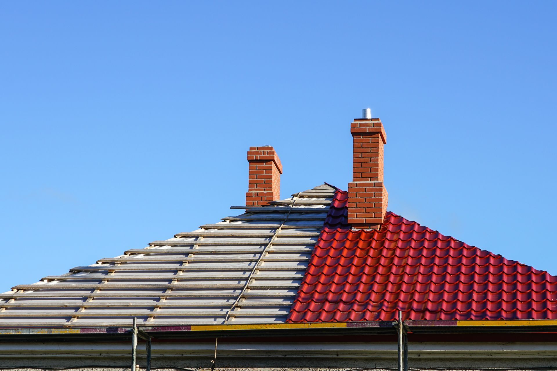 Partially replaced red tile roof with two brick chimneys, against a blue sky.
