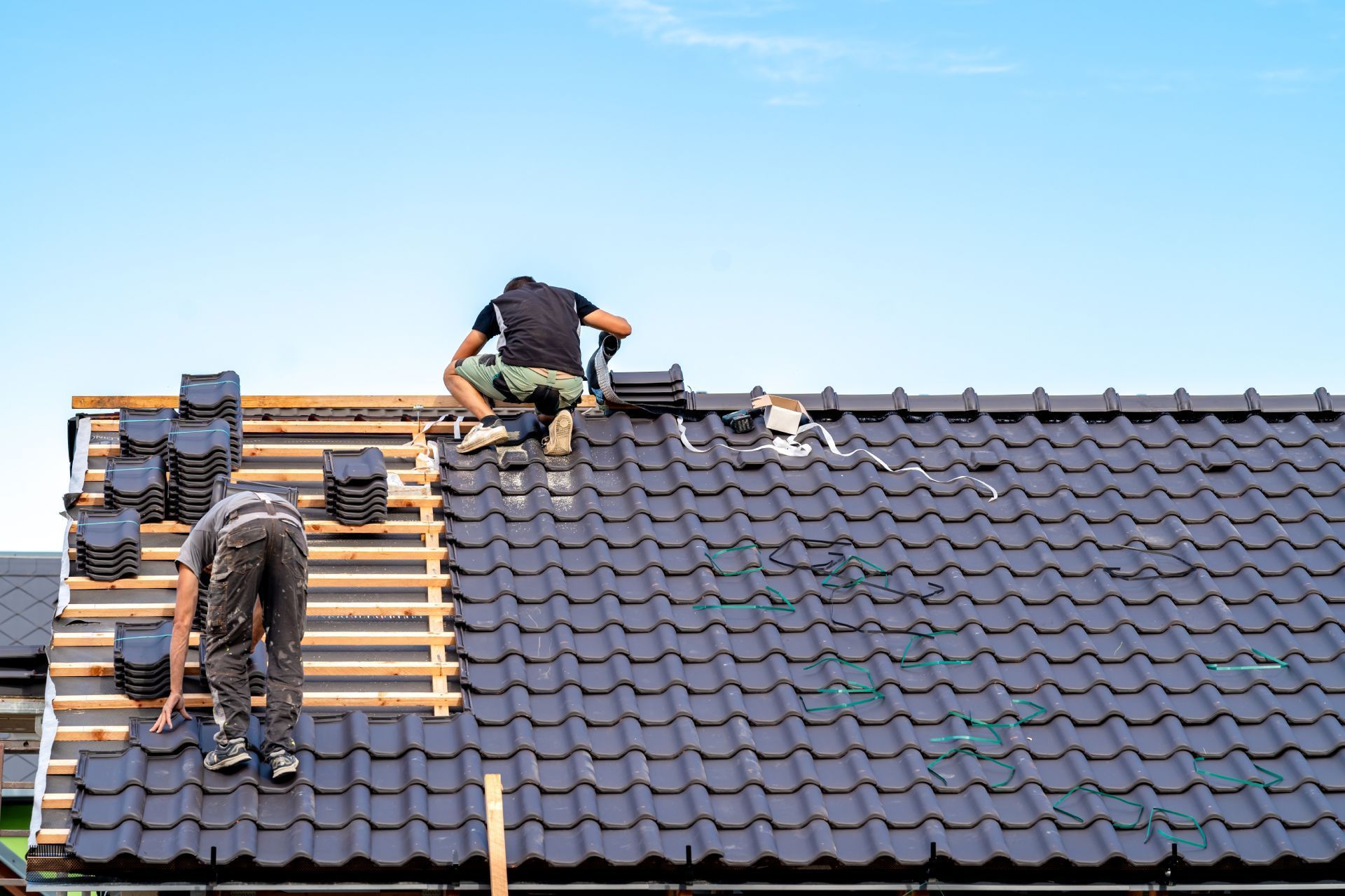 Two roofers installing black tiles on a roof against a blue sky.
