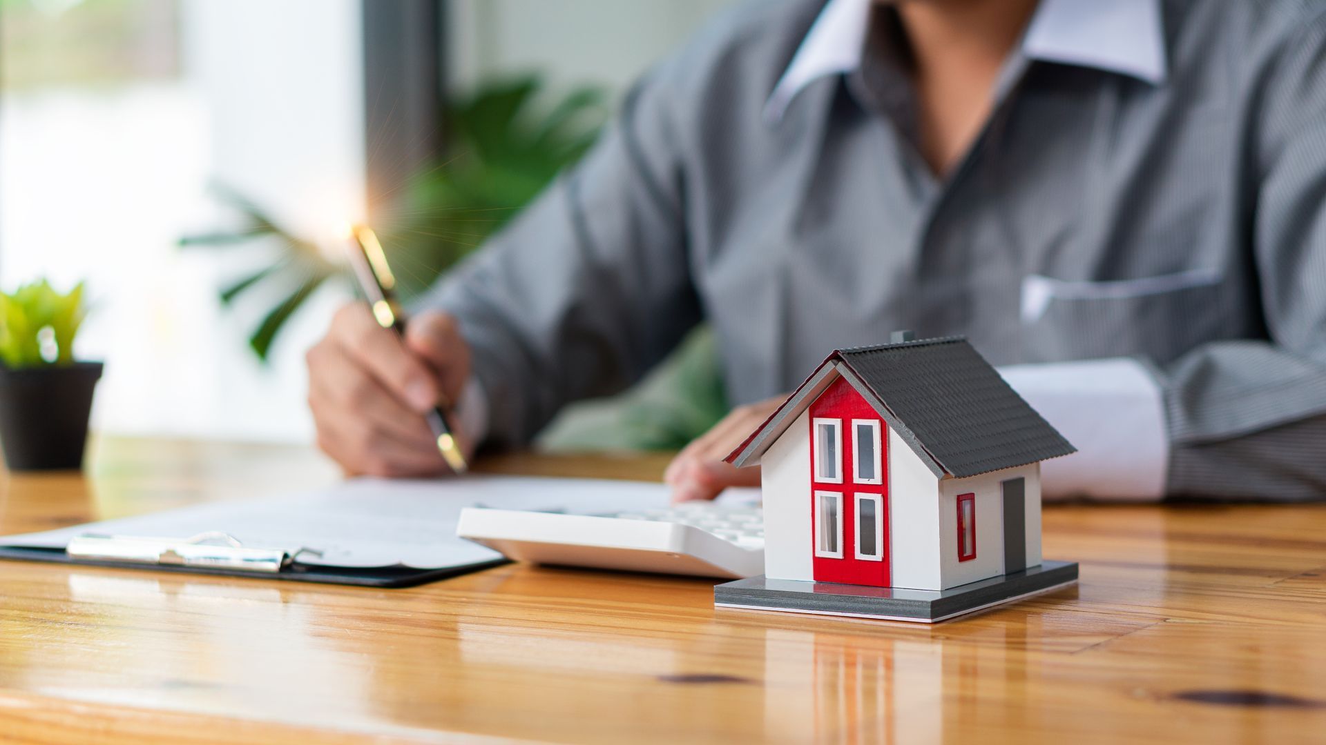 Person using calculator and pen next to a model house on a desk.