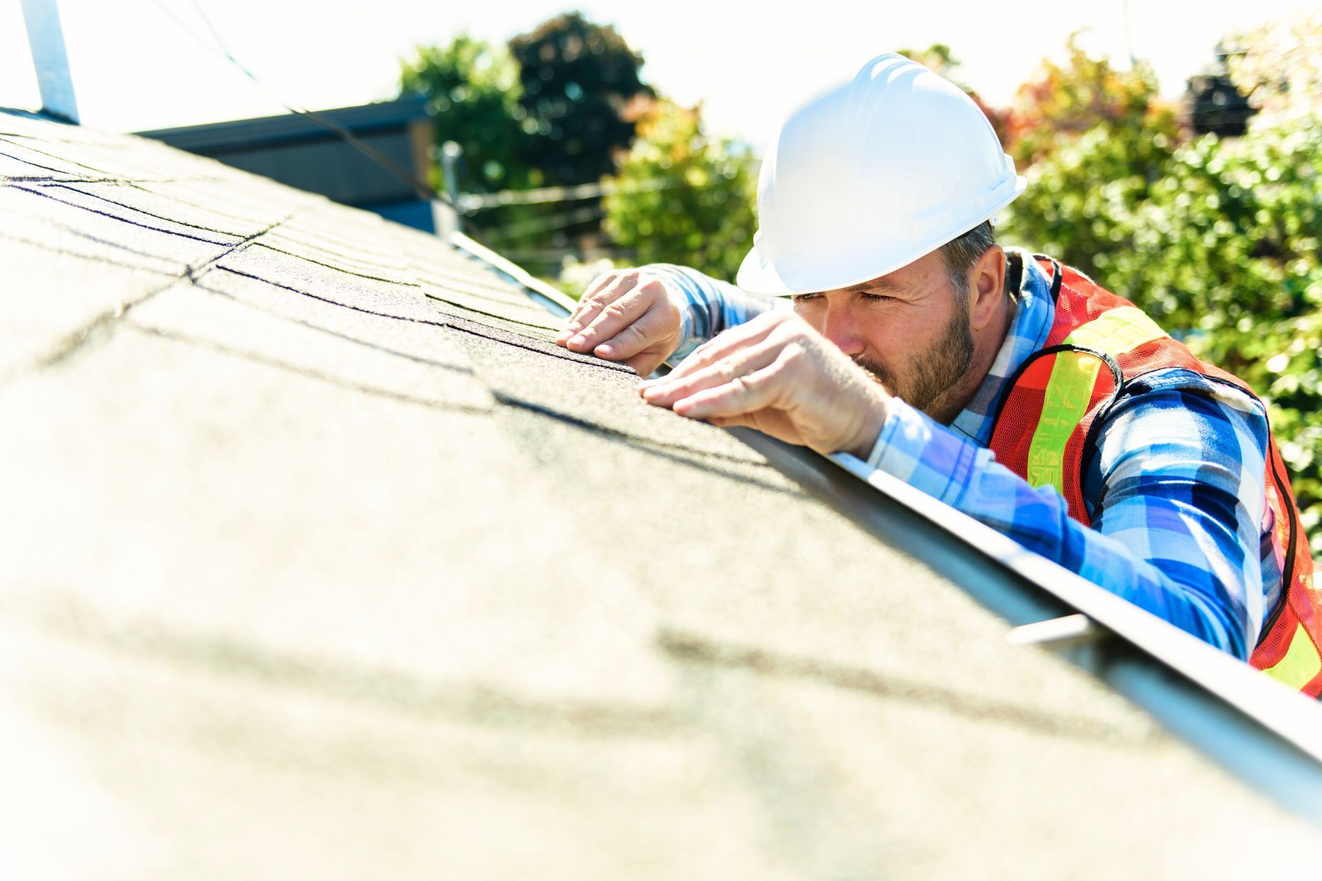 Roofer in white hard hat and safety vest inspecting shingles on a roof.