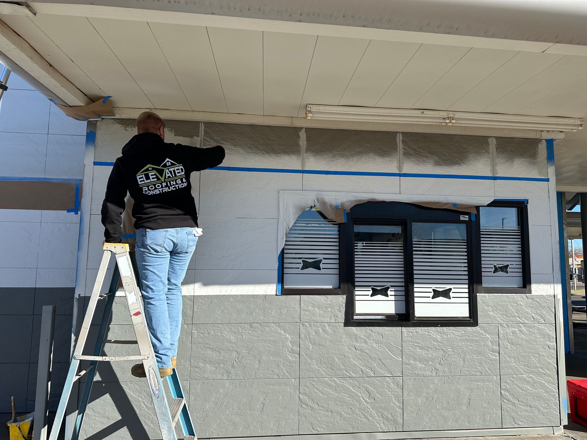 Person on a ladder painting a building's exterior. Blue tape protects windows, gray and white paint visible.
