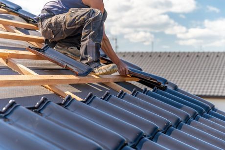 Roofer installing dark gray roof tiles on a wooden frame under a partly cloudy sky.