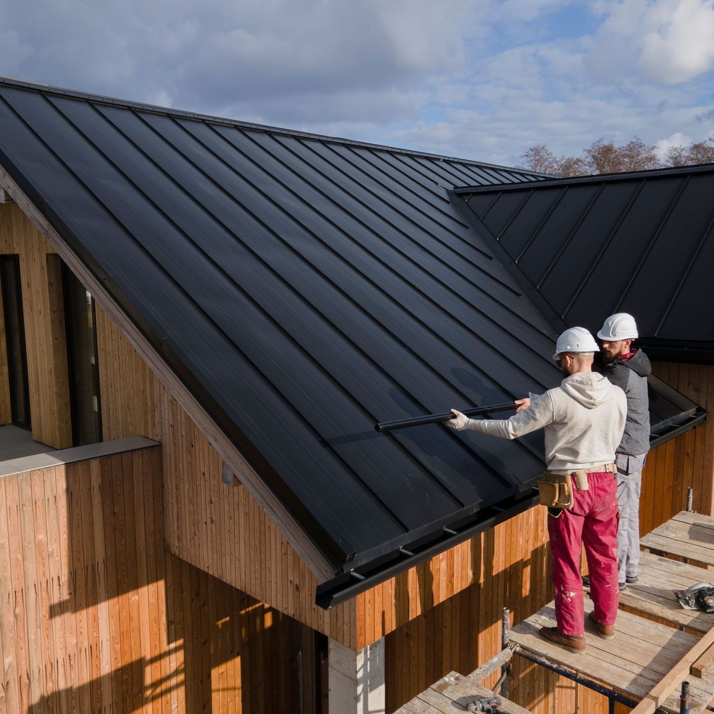 Two construction workers on a roof installing black metal panels on a wooden house.