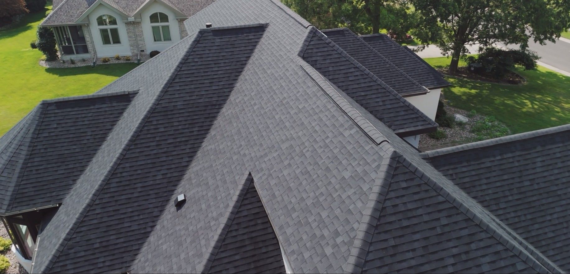 View of a dark gray asphalt shingle roof on a multi-sectioned house. Green lawn and trees are visible.