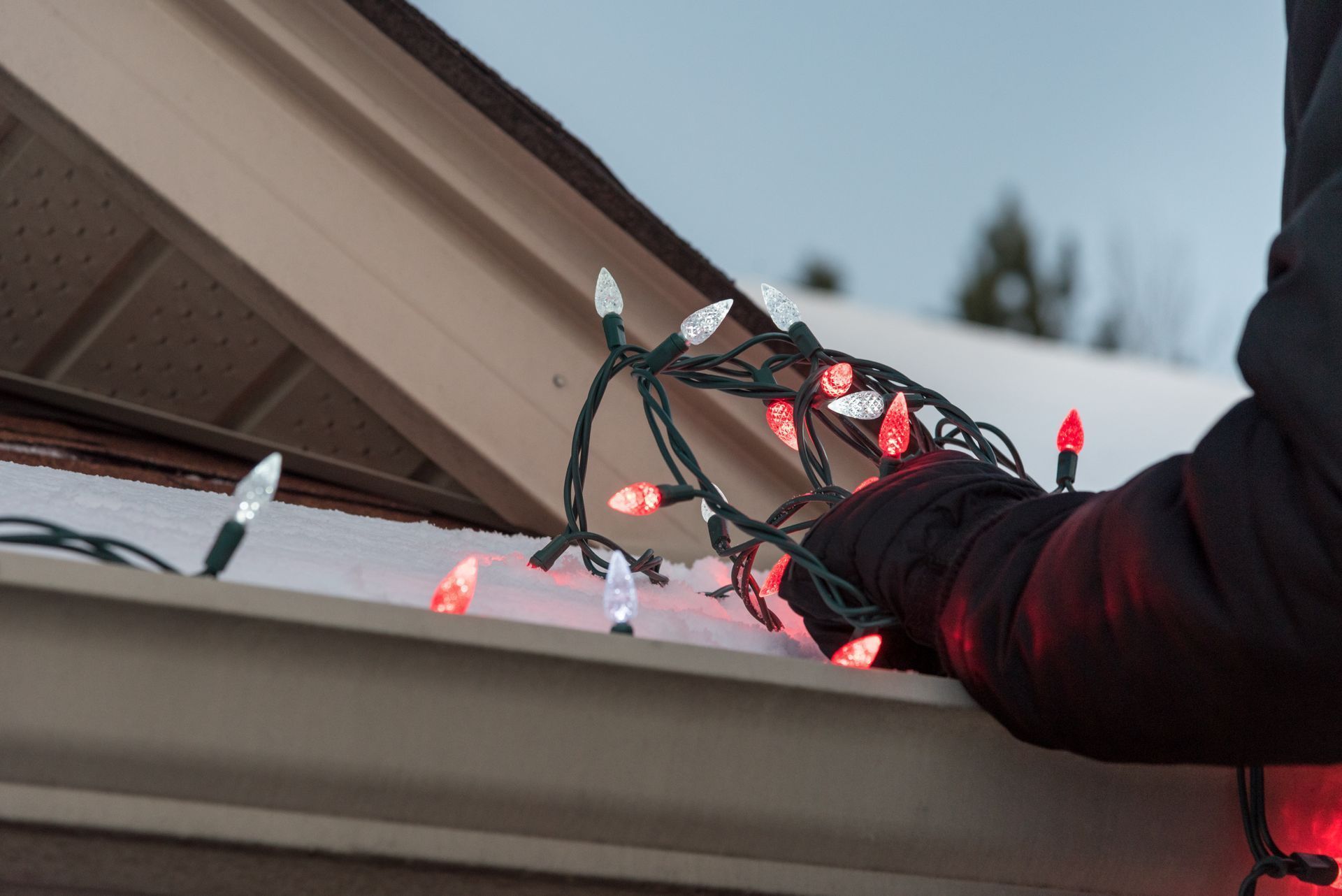Person hangs red and white Christmas lights on a snowy roof gutter.