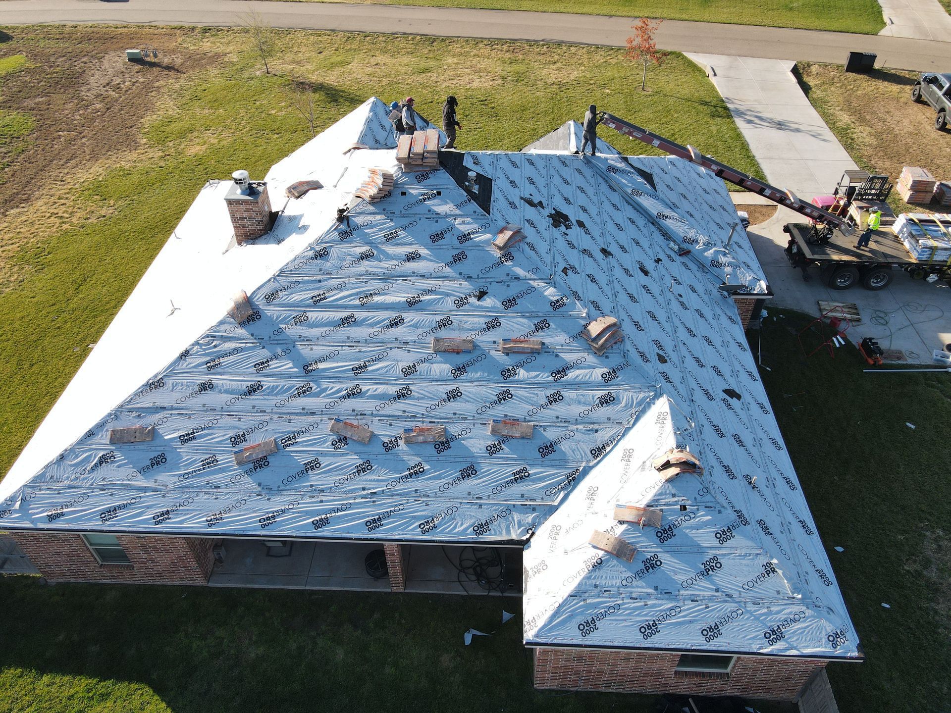 A house with roofing under construction. White underlayment covers the roof, with workers present. A crane is also visible.