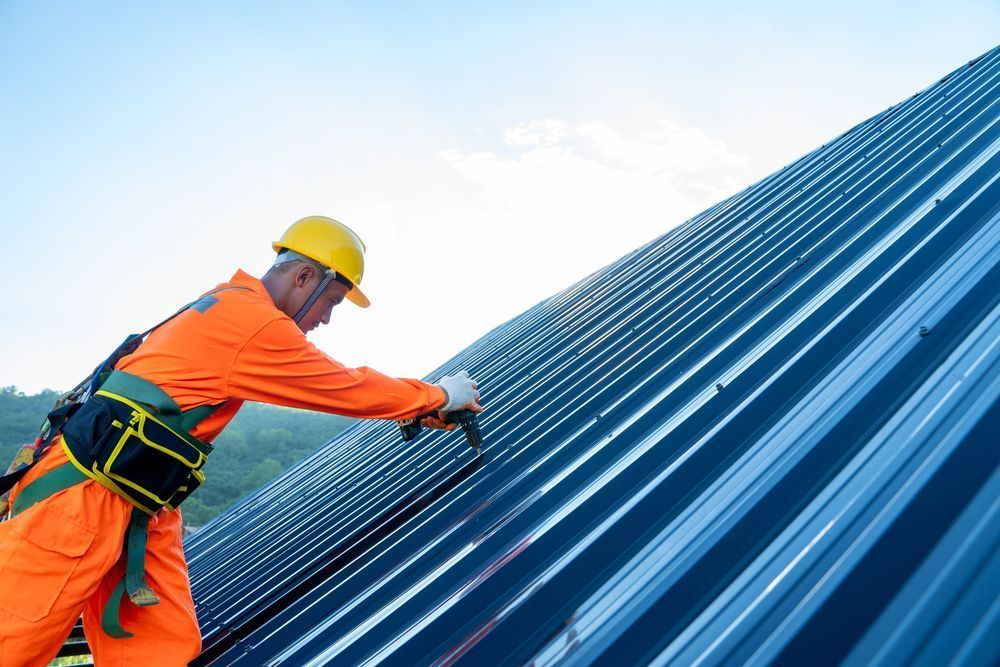 Roofer in orange jumpsuit and helmet working on a metal roof, securing panels with a tool.