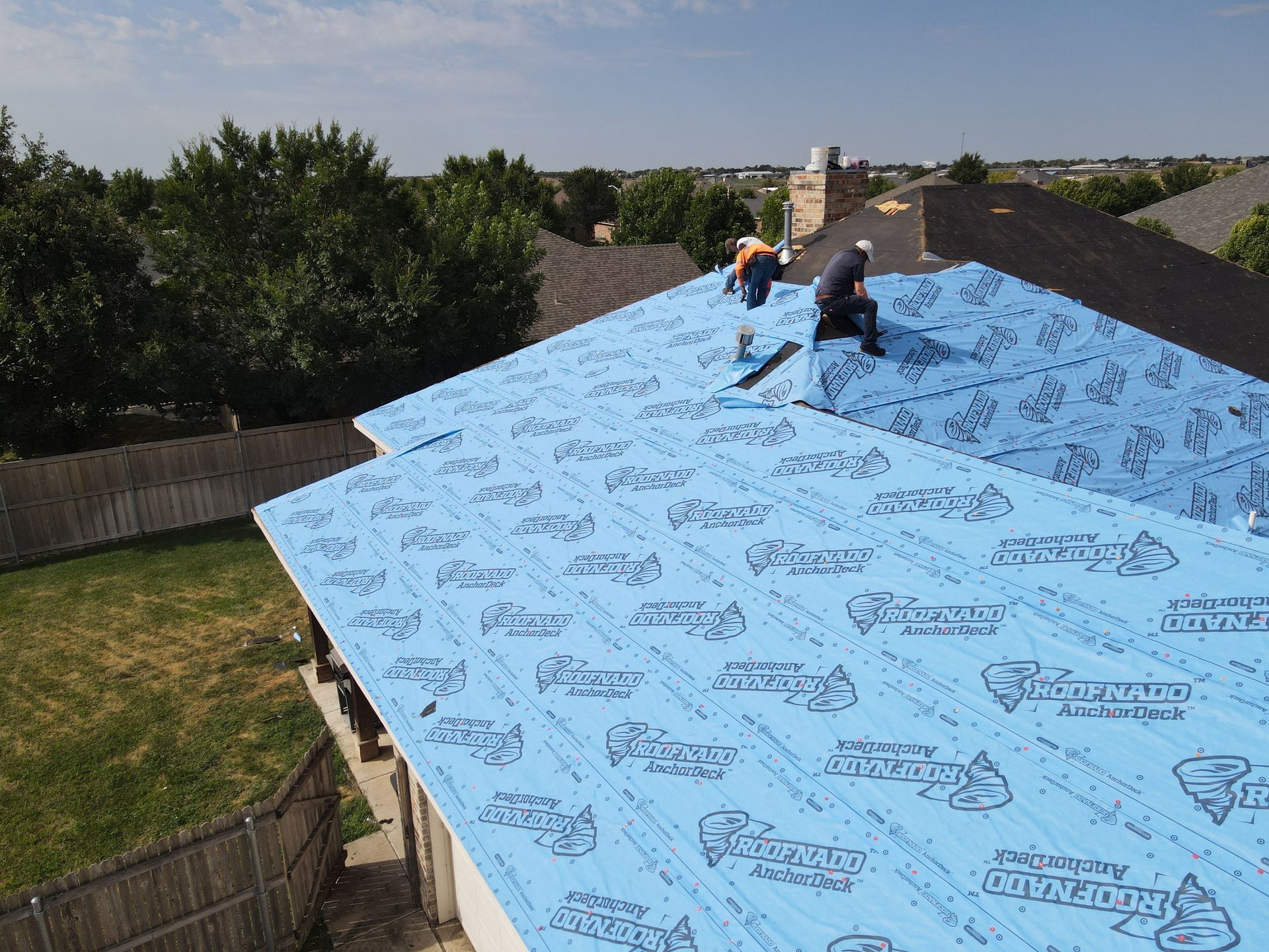 Two workers on a roof covered in blue underlayment, preparing for new shingles.