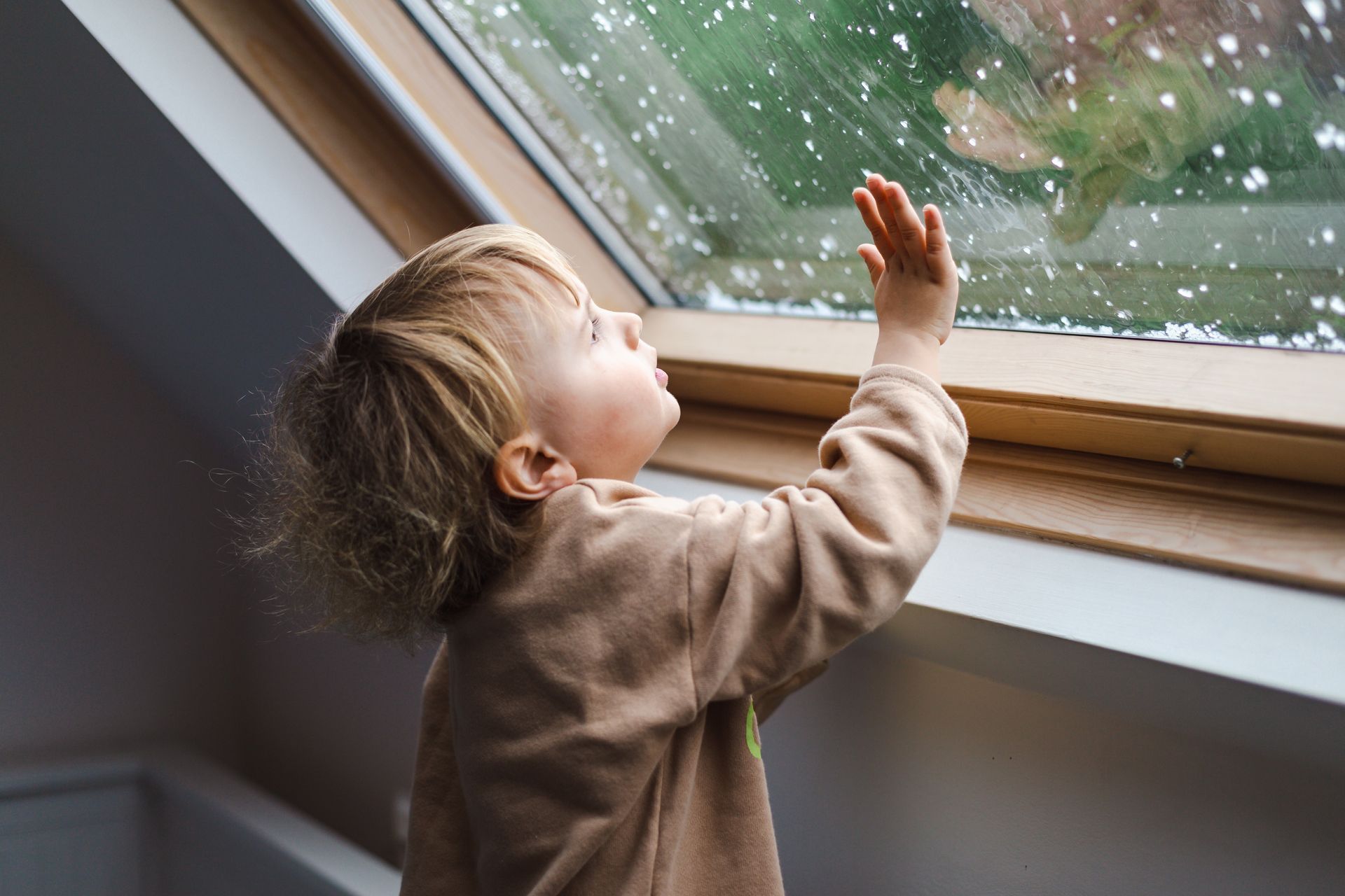 Child looking up, touching a rain-streaked window. Blonde hair, brown shirt, indoors, overcast day.