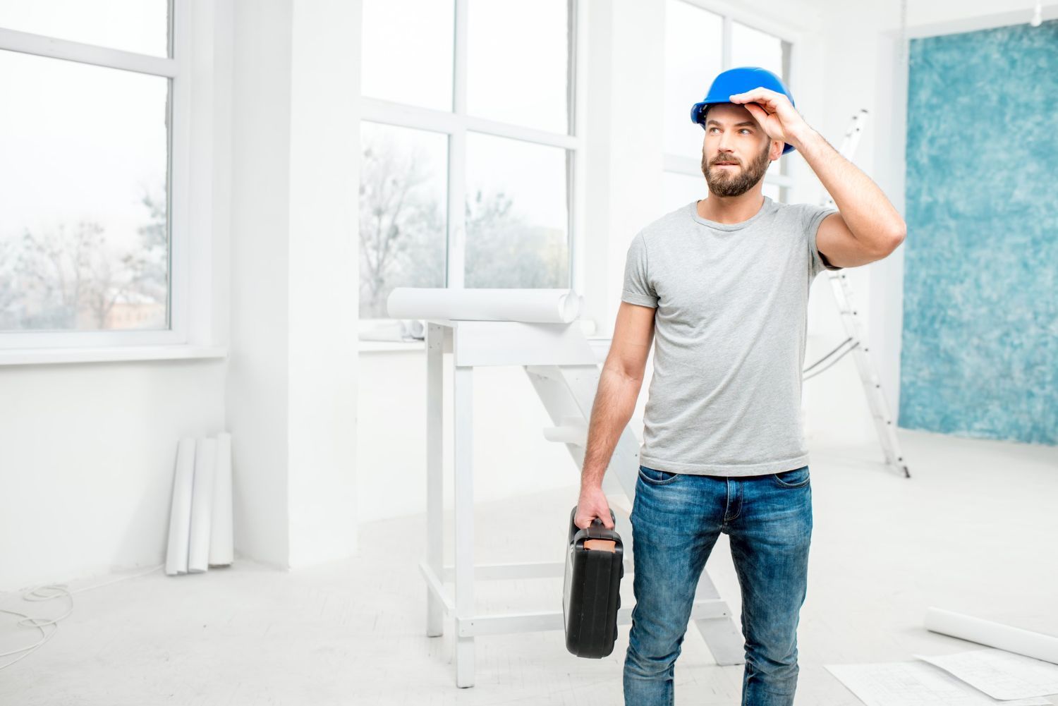 Construction worker wearing a blue hard hat, holding a toolbox, and looking up in a room.