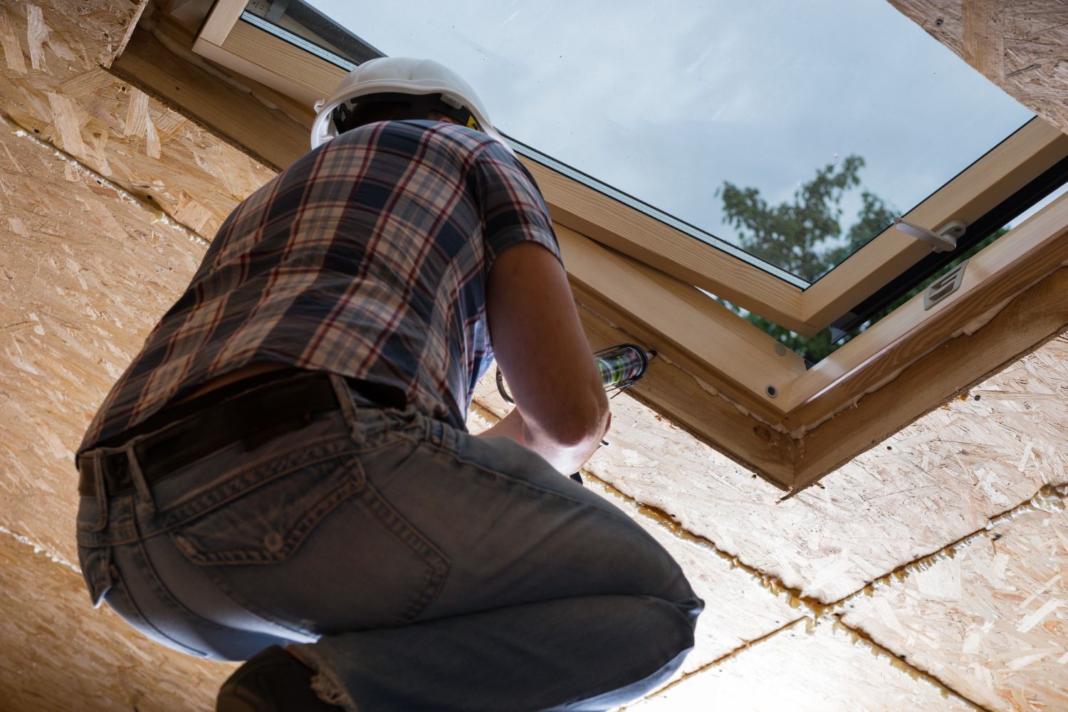 Person in plaid shirt and jeans installing a skylight, viewed from below.