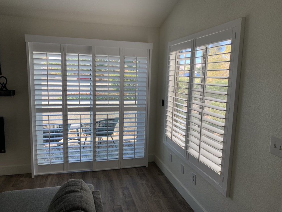White window shutters in a room with hardwood flooring, covering a sliding glass door and a window, letting in sunlight.