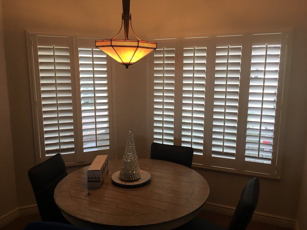 Dining room with a round table and three chairs, a bay window with shutters, and a hanging light.