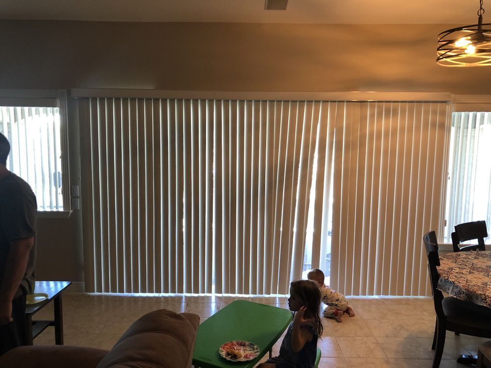 Living room with vertical blinds, table with a child, and another child crawling on the floor.