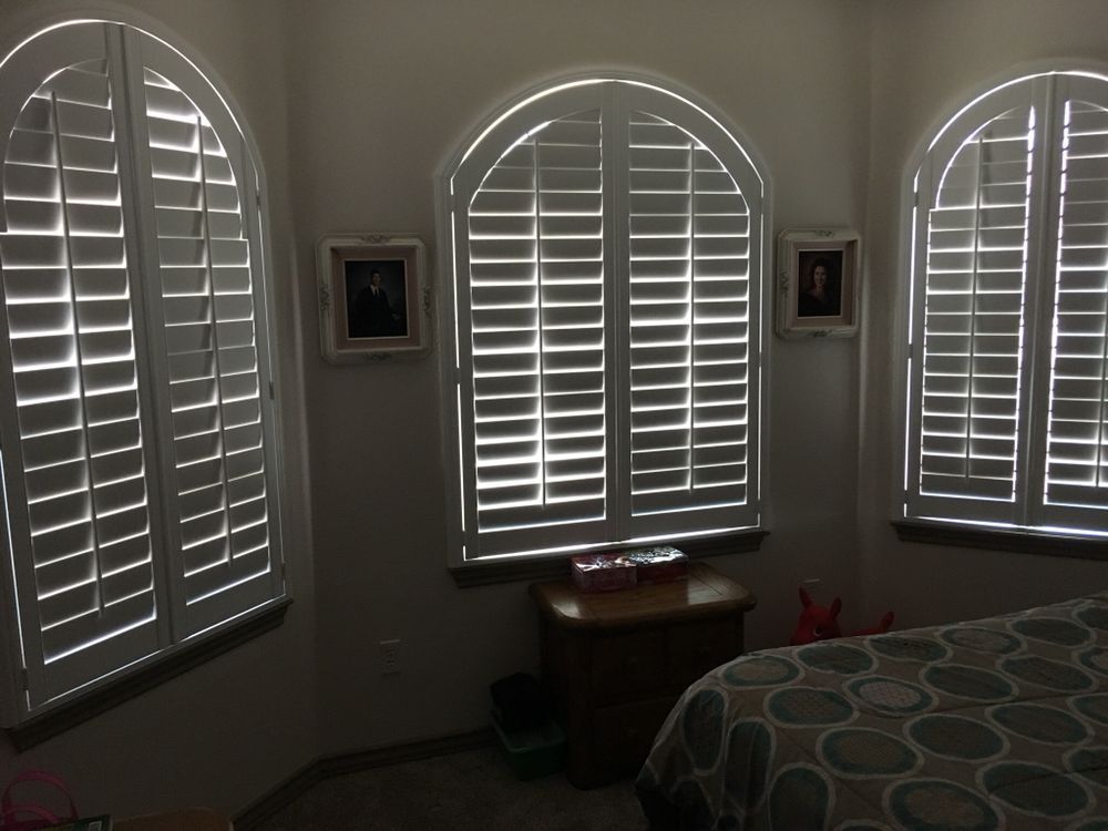 White arched window shutters in a bedroom. Sunlight streams through the slats. There are two framed photos on the wall.