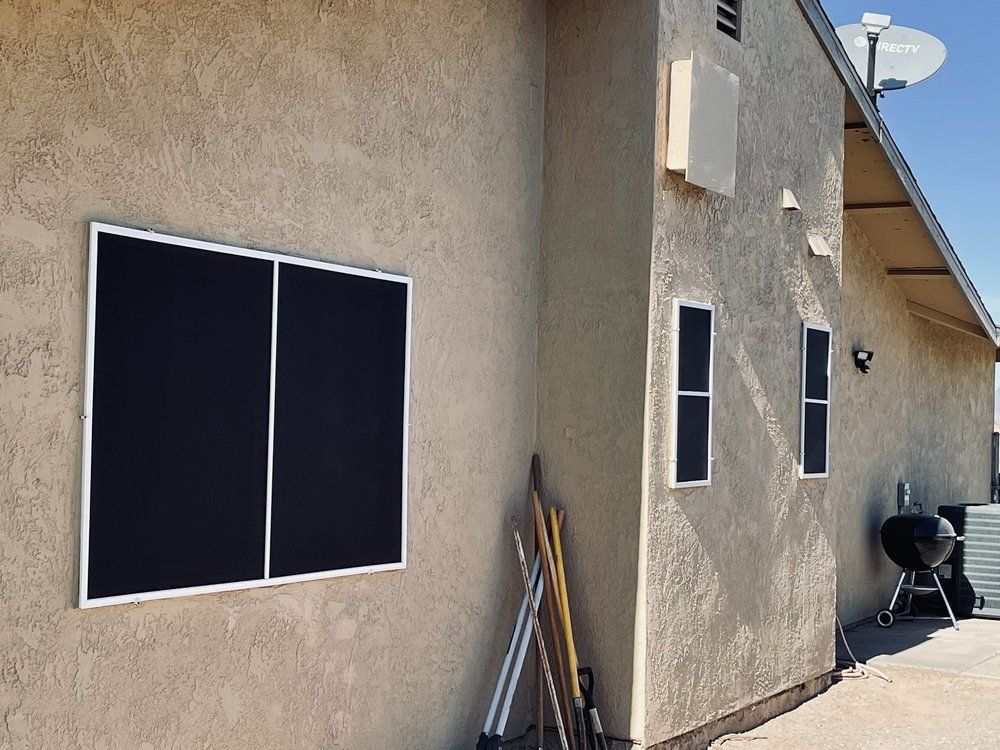 Exterior of a stucco building with multiple windows. Two large, dark screens are visible, along with yard tools and a grill.
