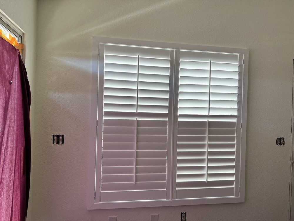 White plantation shutters on a window with sunlight streaming in, against a white wall.