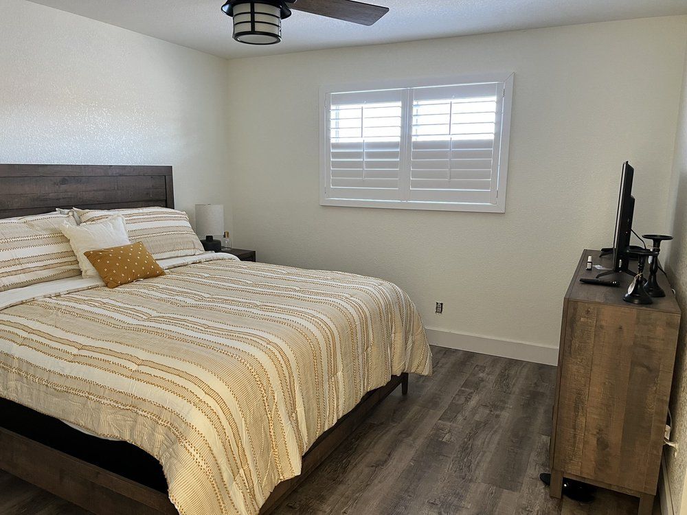 Bedroom with a bed, dresser, window with shutters, and ceiling fan. Warm tones, light walls, and wood-look flooring.