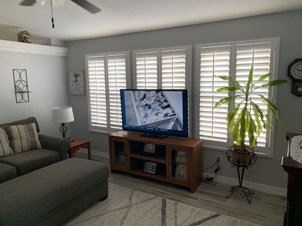Living room with gray sofa, TV on wooden console, and large windows with shutters.