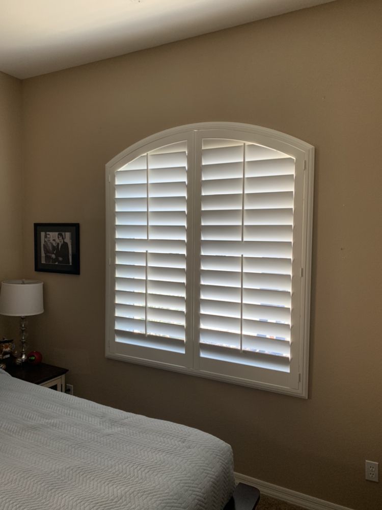 Bedroom with arched window covered by white shutters; tan walls, bedside table, lamp, and bed.