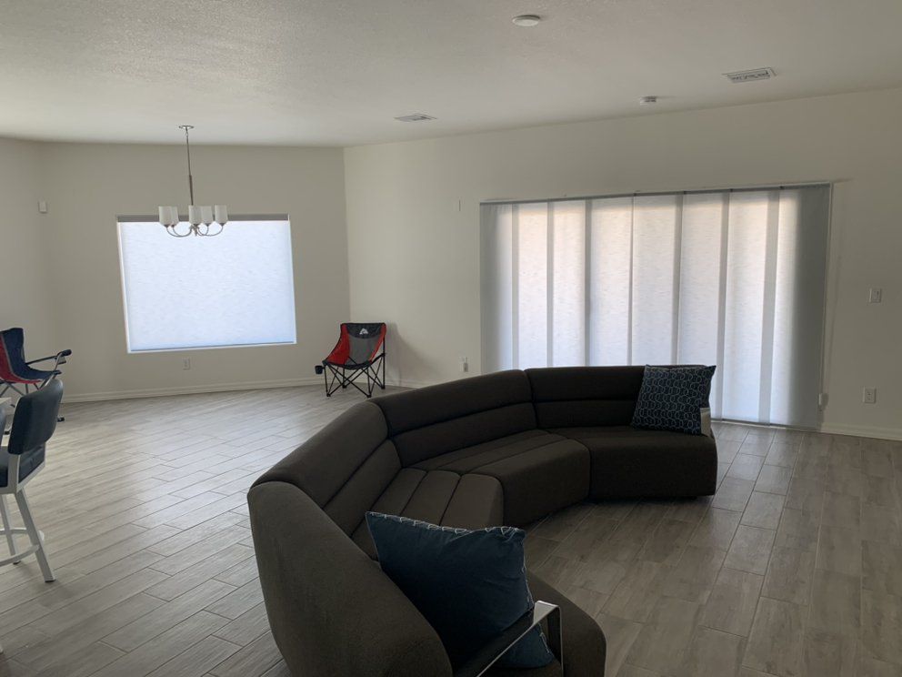 Spacious living room with a curved brown sectional sofa, large windows with white blinds, and light-colored flooring.