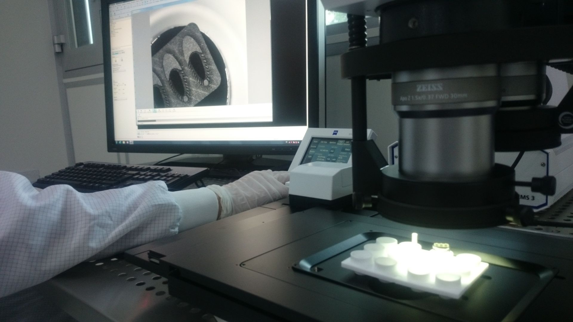 A person in a lab coat uses a microscope to examine small, bright objects, with a computer screen showing a close-up view of them.