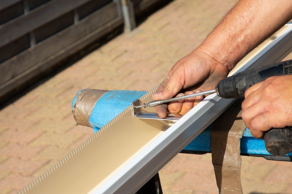 Hands using a drill to attach a metal piece to a tan gutter; outdoor setting.