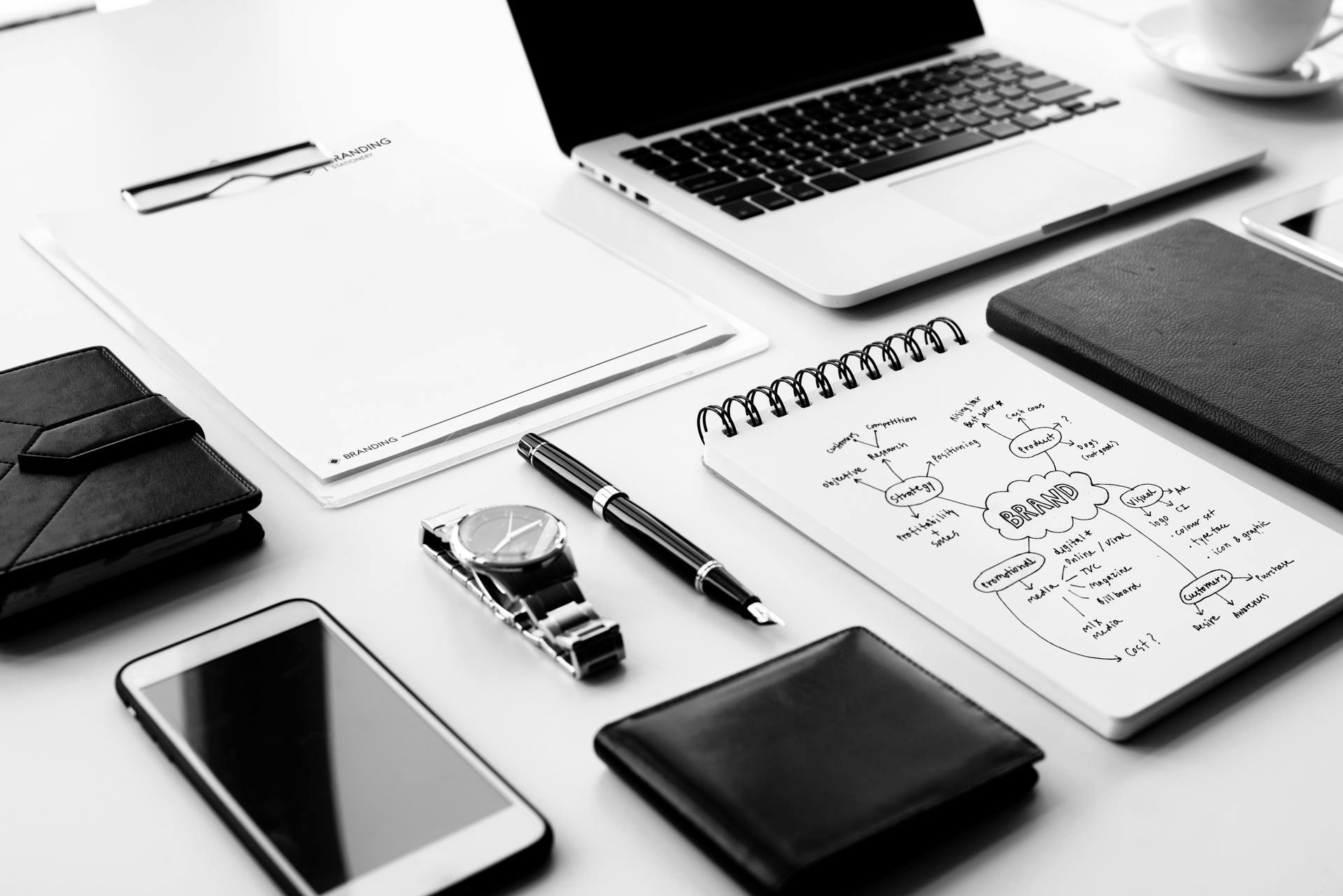 Desk with laptop, notebook, phone, watch, pen, wallets, and clipboard.