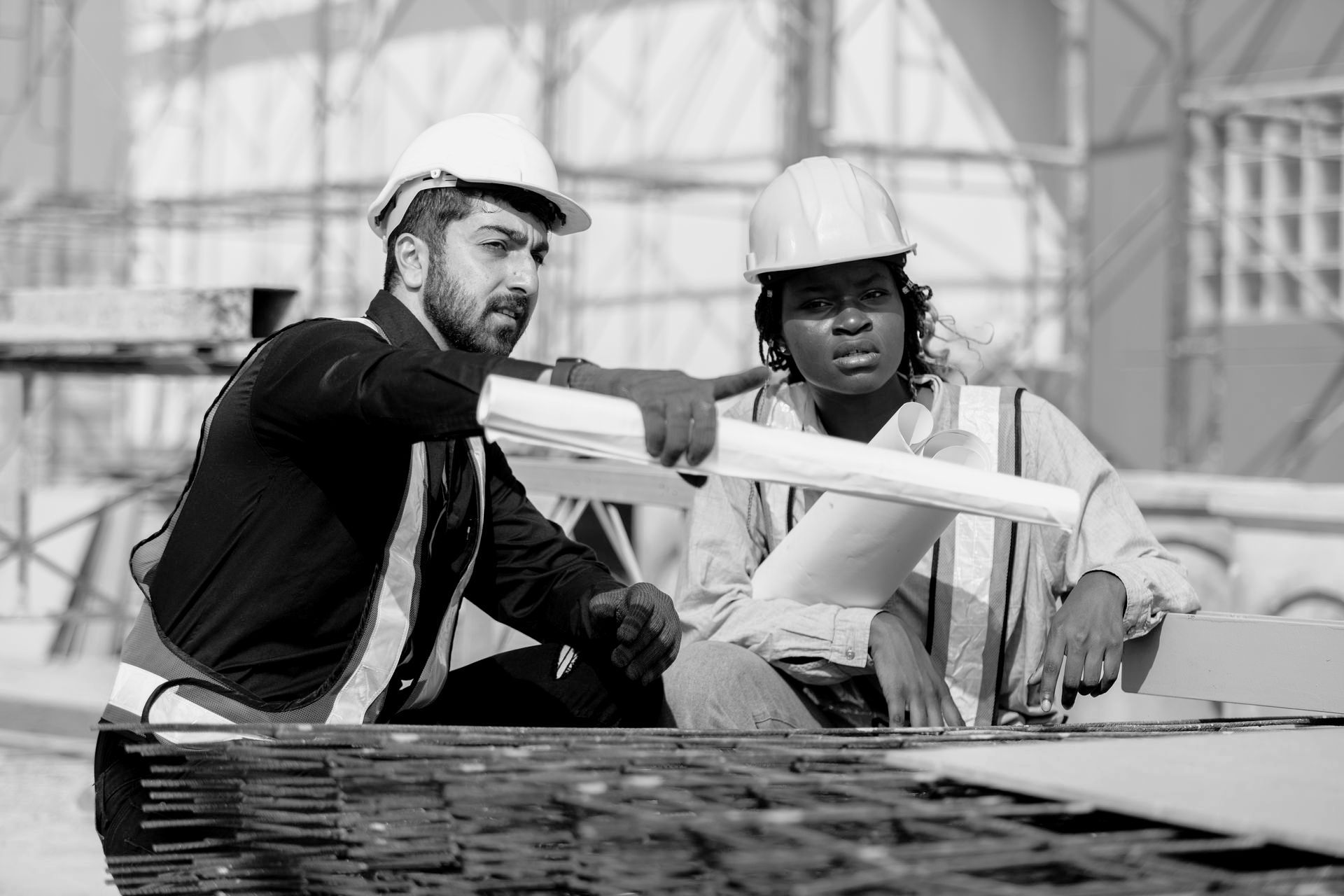 Two construction workers, a man pointing at blueprints, a woman looking on; outdoors.