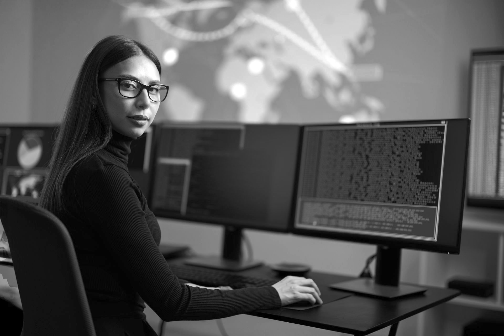 Woman with glasses at computer in a control room; world map and data on monitors in the background.