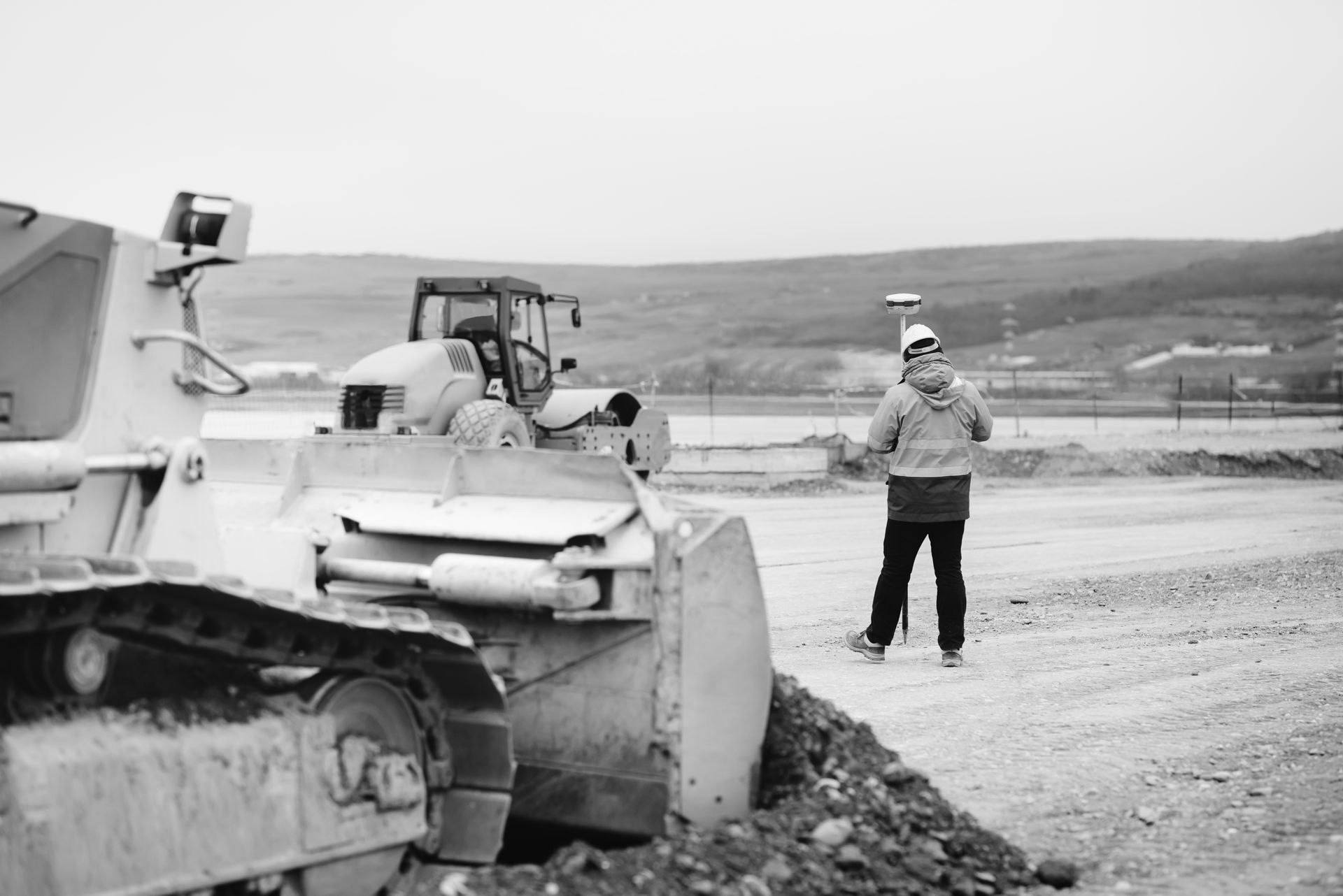 Man surveying construction site with bulldozer and tractor in the background, overcast day.