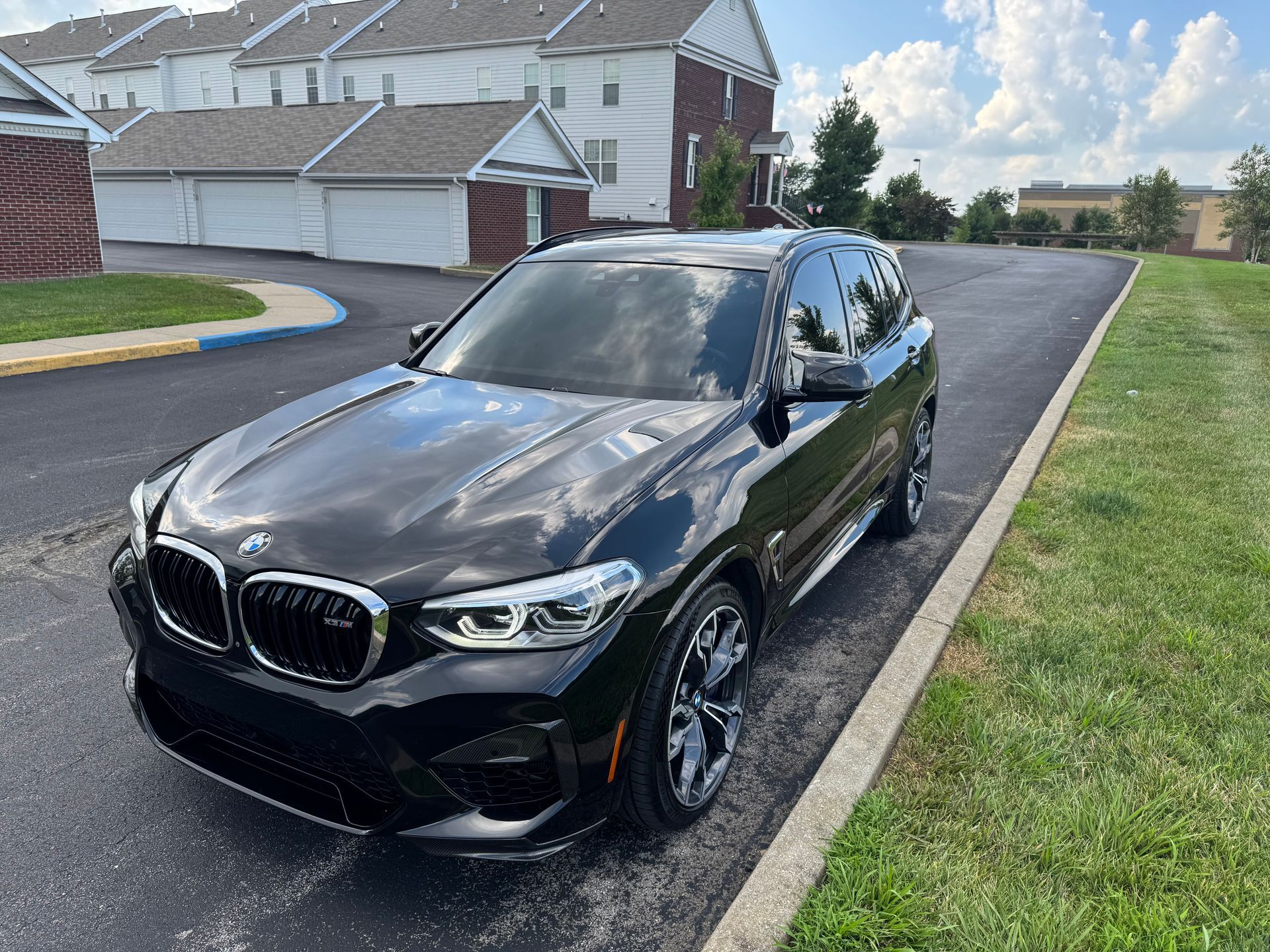 Black BMW X3 M SUV parked on asphalt next to grass, with apartment buildings in the background.