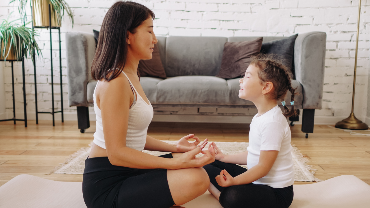 A woman and a little girl are sitting on a yoga mat in a living room.