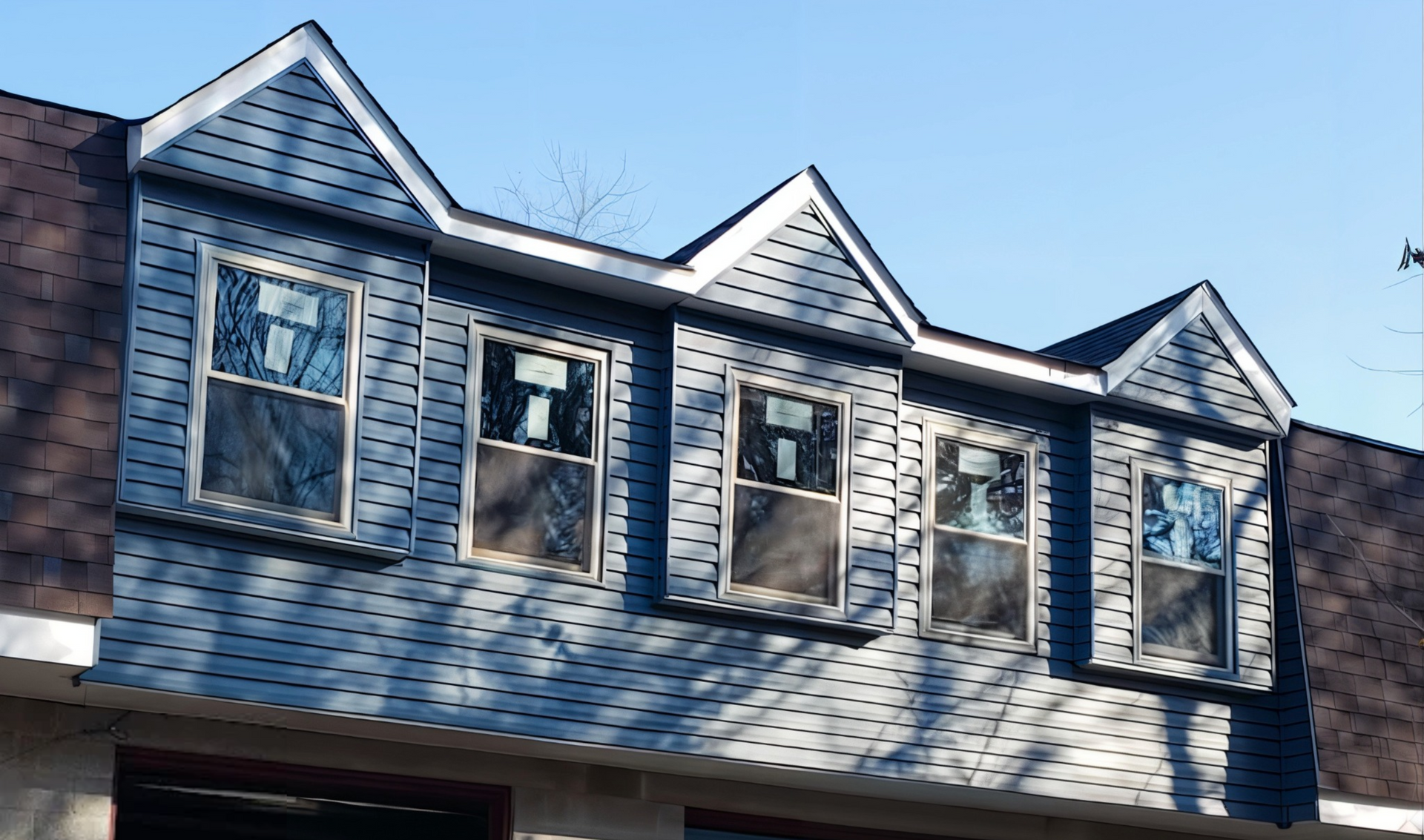 Three dormers with windows on a house, siding is blue. The roof is brown and the sky is clear.