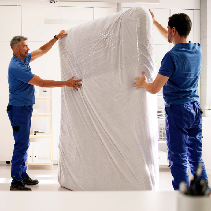 Two men are wrapping a mattress in plastic wrap.