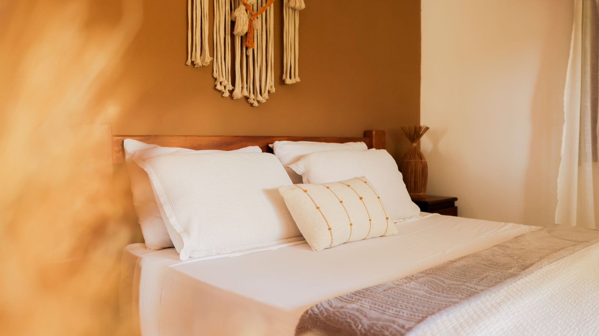 Quarto com cabeceira de madeira, roupa de cama branca e painel de macramê na parede.