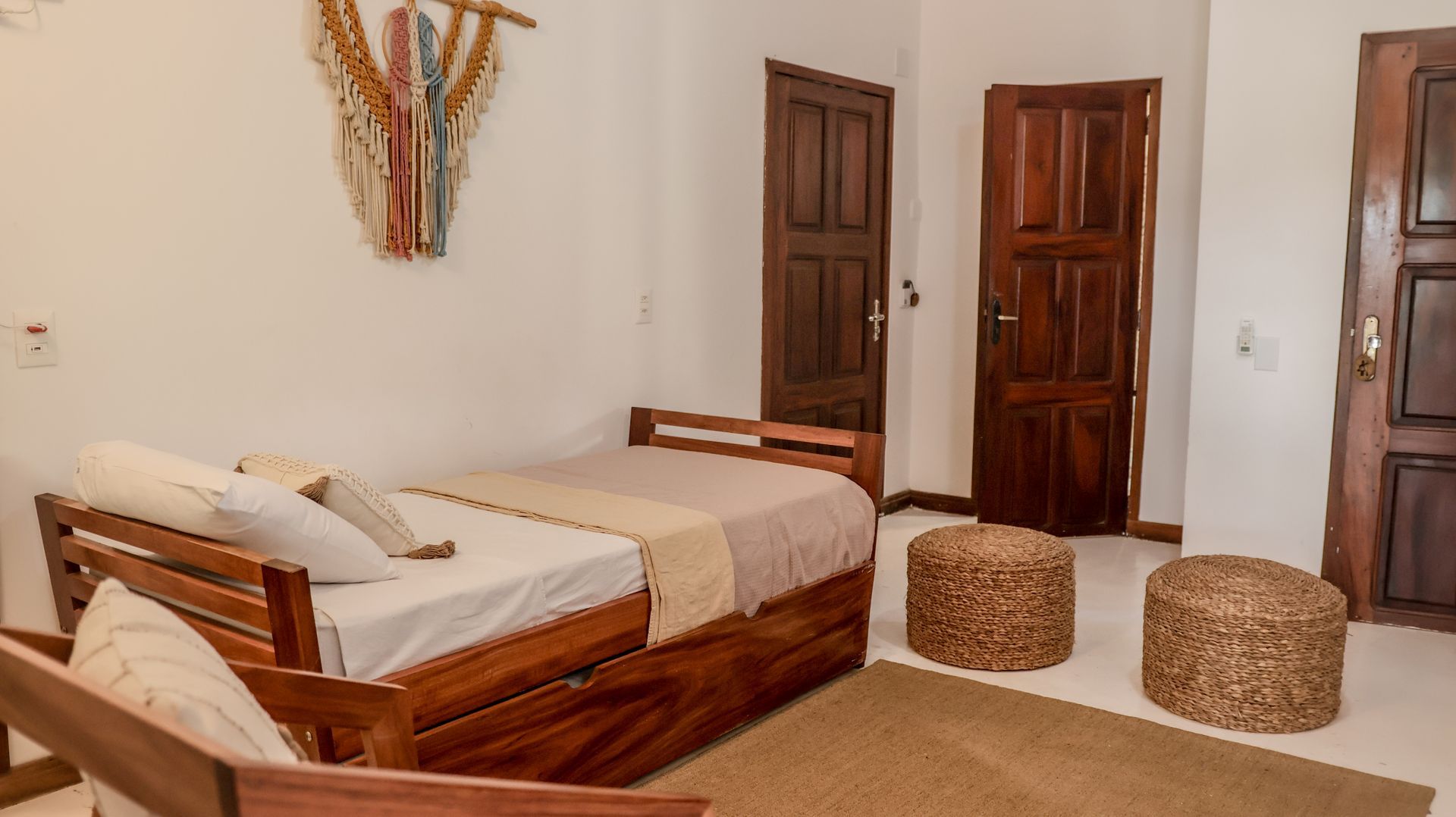 Quarto com divã de madeira, roupa de cama em tons neutros, pufes de vime e portas de madeira.