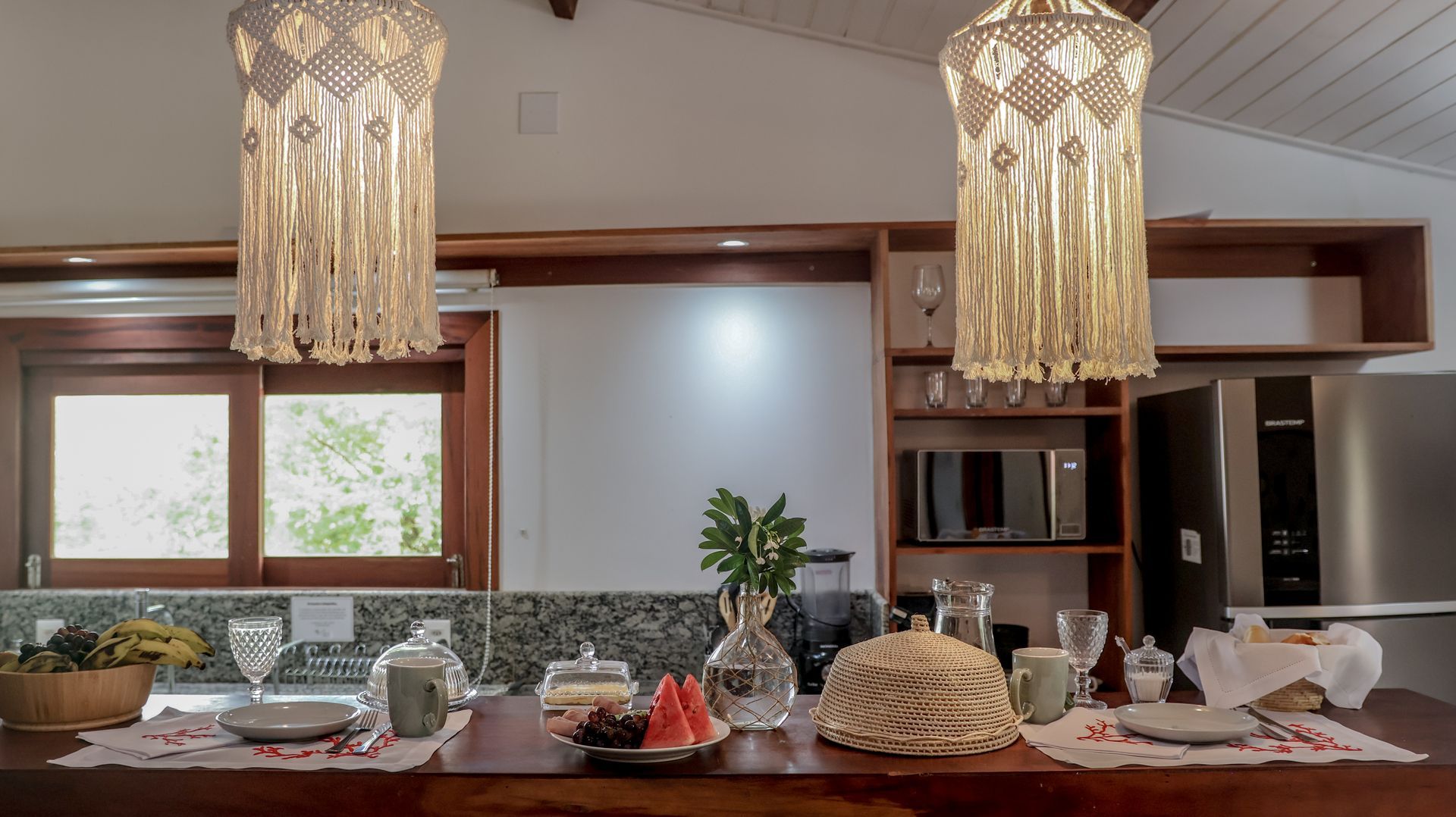 Balcão de cozinha com luminárias de macramê, pronto para uma refeição. Inclui copos, alimentos e eletrodomésticos embutidos.