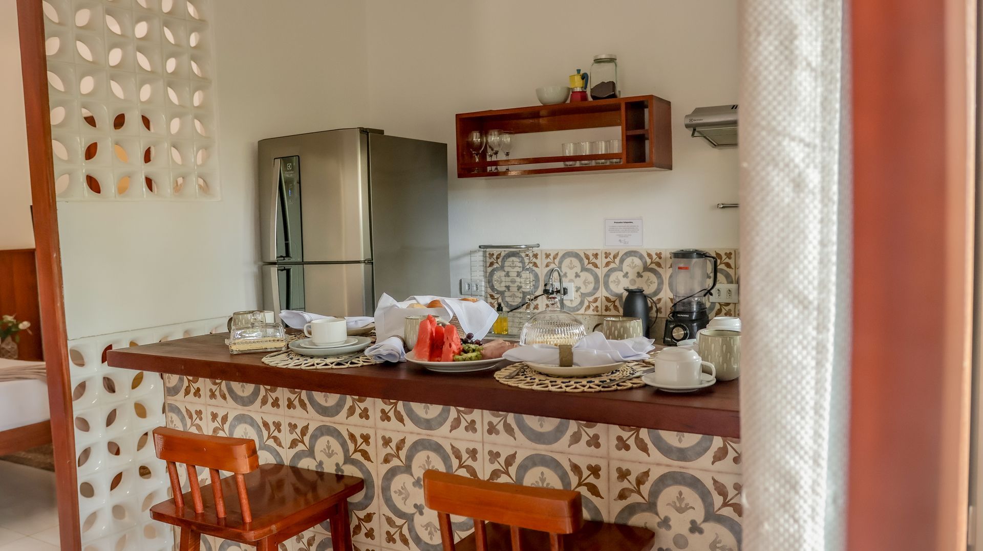 Bancada da cozinha com frutas, louças e banquetas. Geladeira e backsplash de azulejos estampados. Detalhes em madeira.