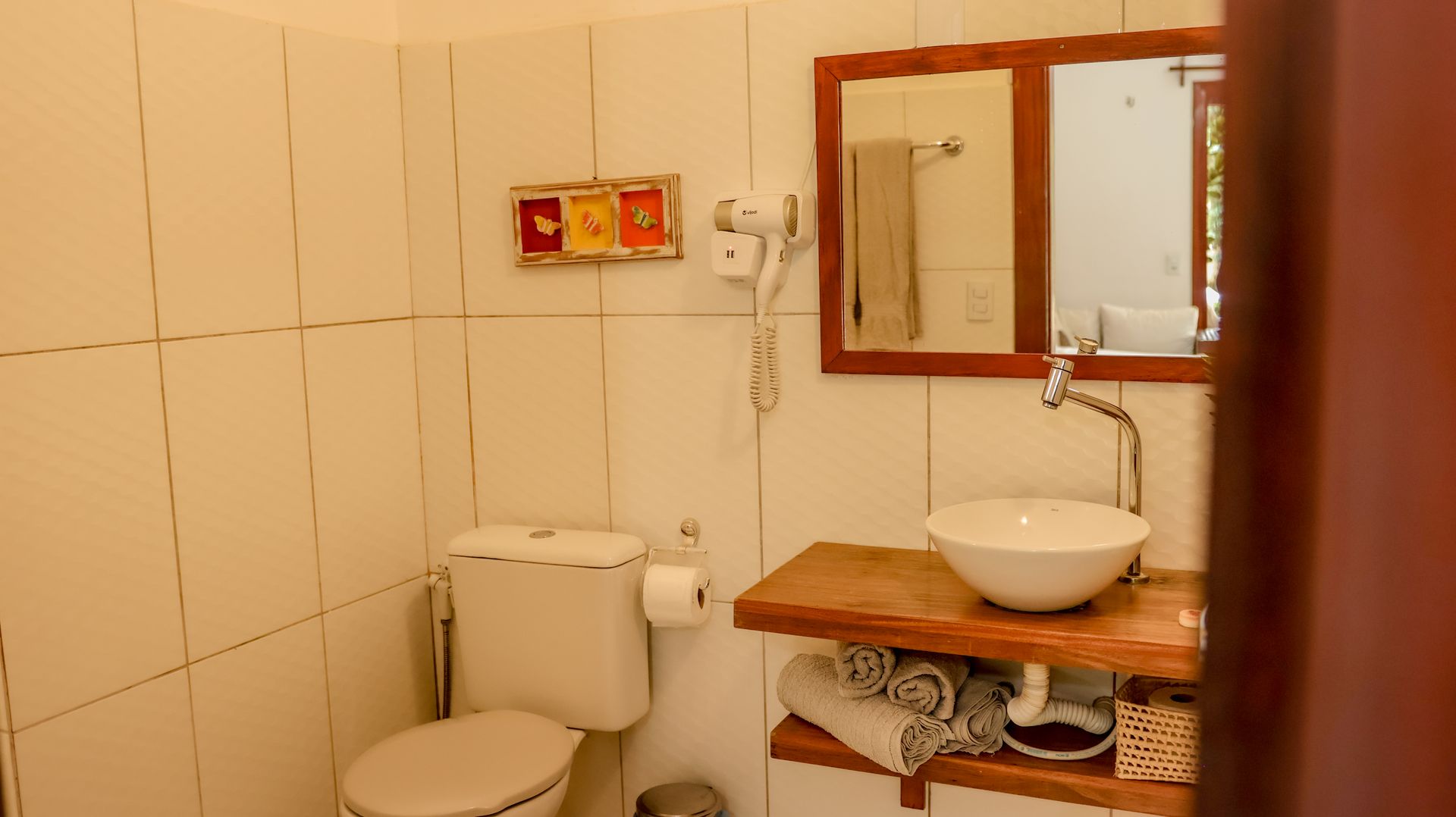 Banheiro com vaso sanitário, pia sobre prateleira de madeira e espelho de parede. Azulejos e decoração brancos.