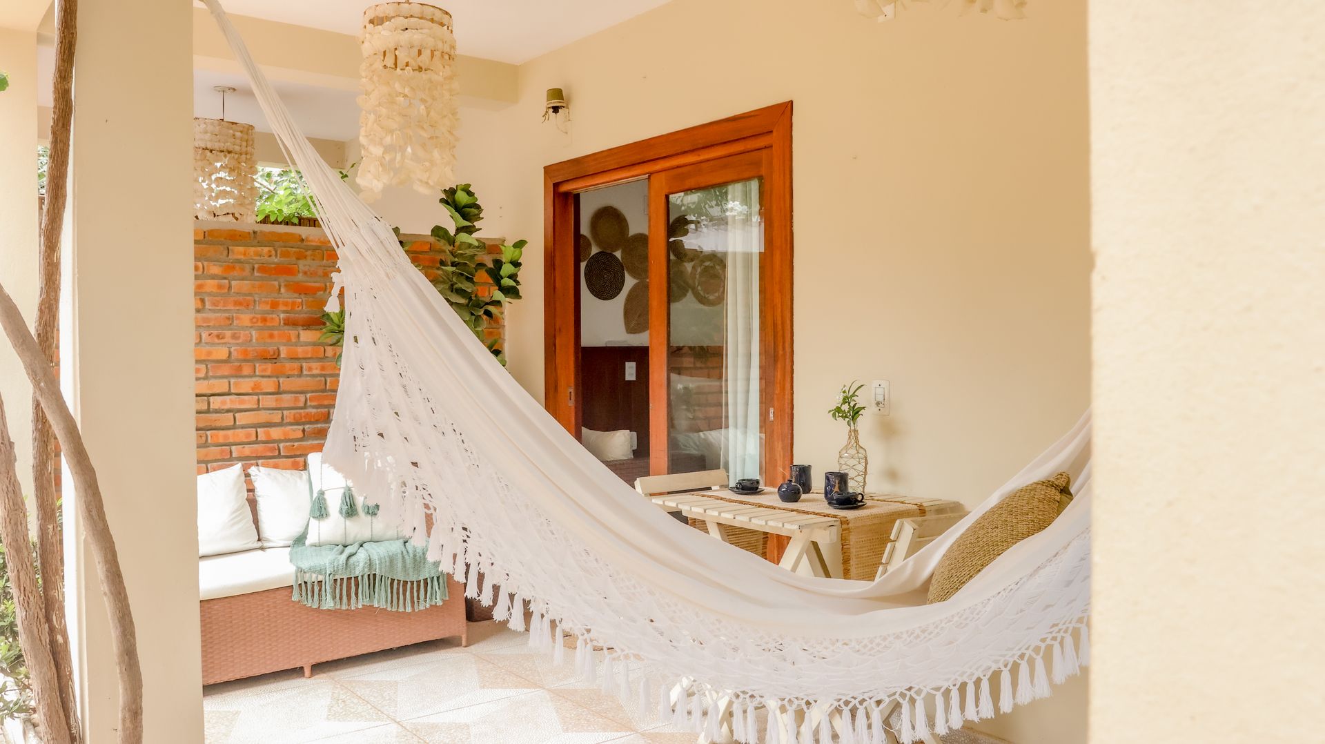 Rede branca em uma varanda com mesa, porta de correr, parede de tijolos e decoração pendurada.