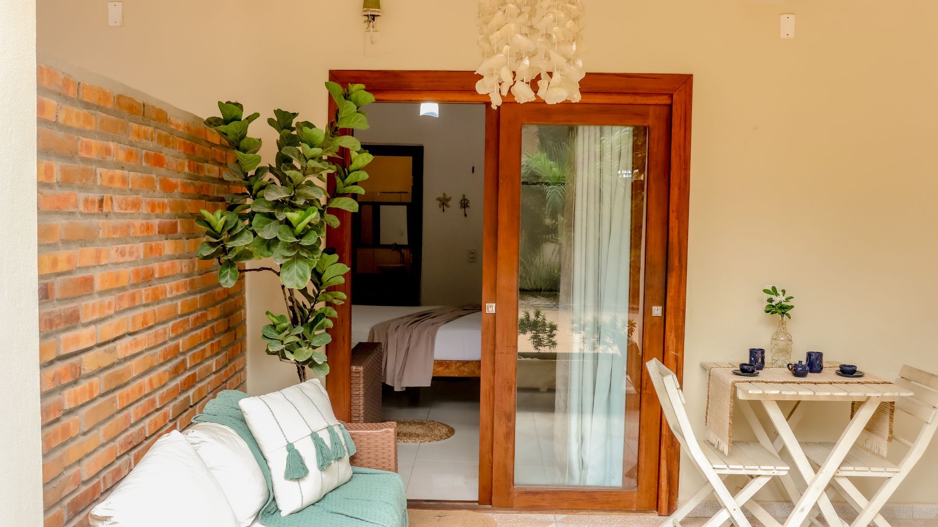 Varanda com sofá azul-petróleo, parede de tijolos e mesa. Portas de madeira dão acesso a um quarto com cama e decoração.