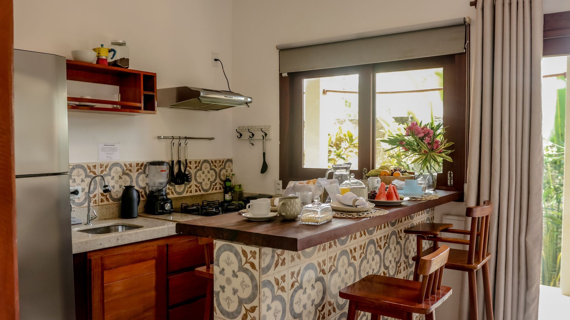 Cozinha com bancada para refeições rápidas, backsplash de azulejos e iluminação natural proveniente de uma janela. Inclui geladeira, fogão e prateleiras abertas.