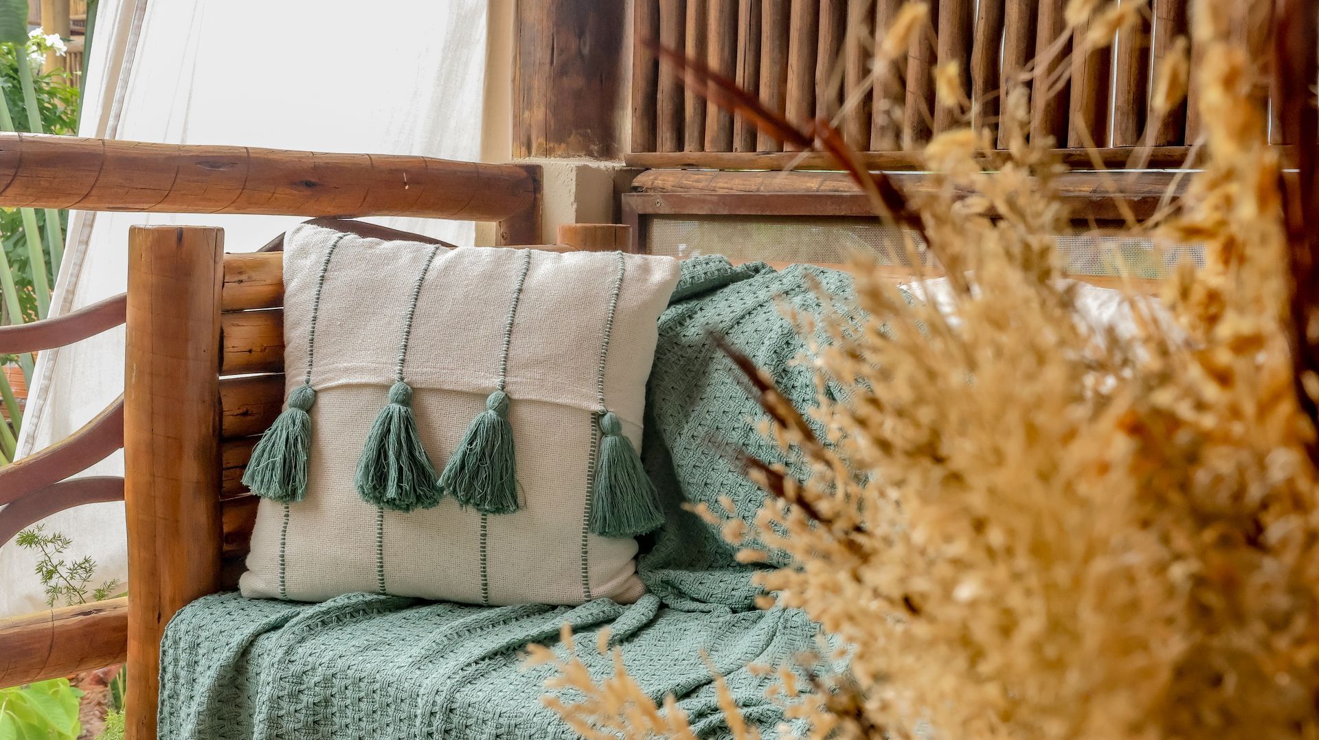 Almofada com borlas verdes sobre uma manta verde, em um banco de madeira, com plantas secas.