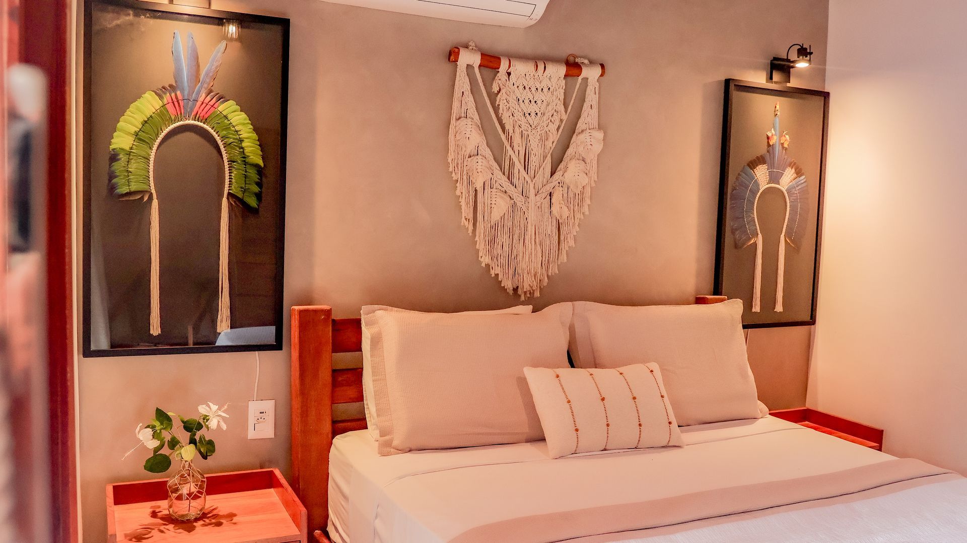 Quarto com obras de arte, painel de macramê na parede e estrutura de cama de madeira. Tons de rosa claro e cinza.