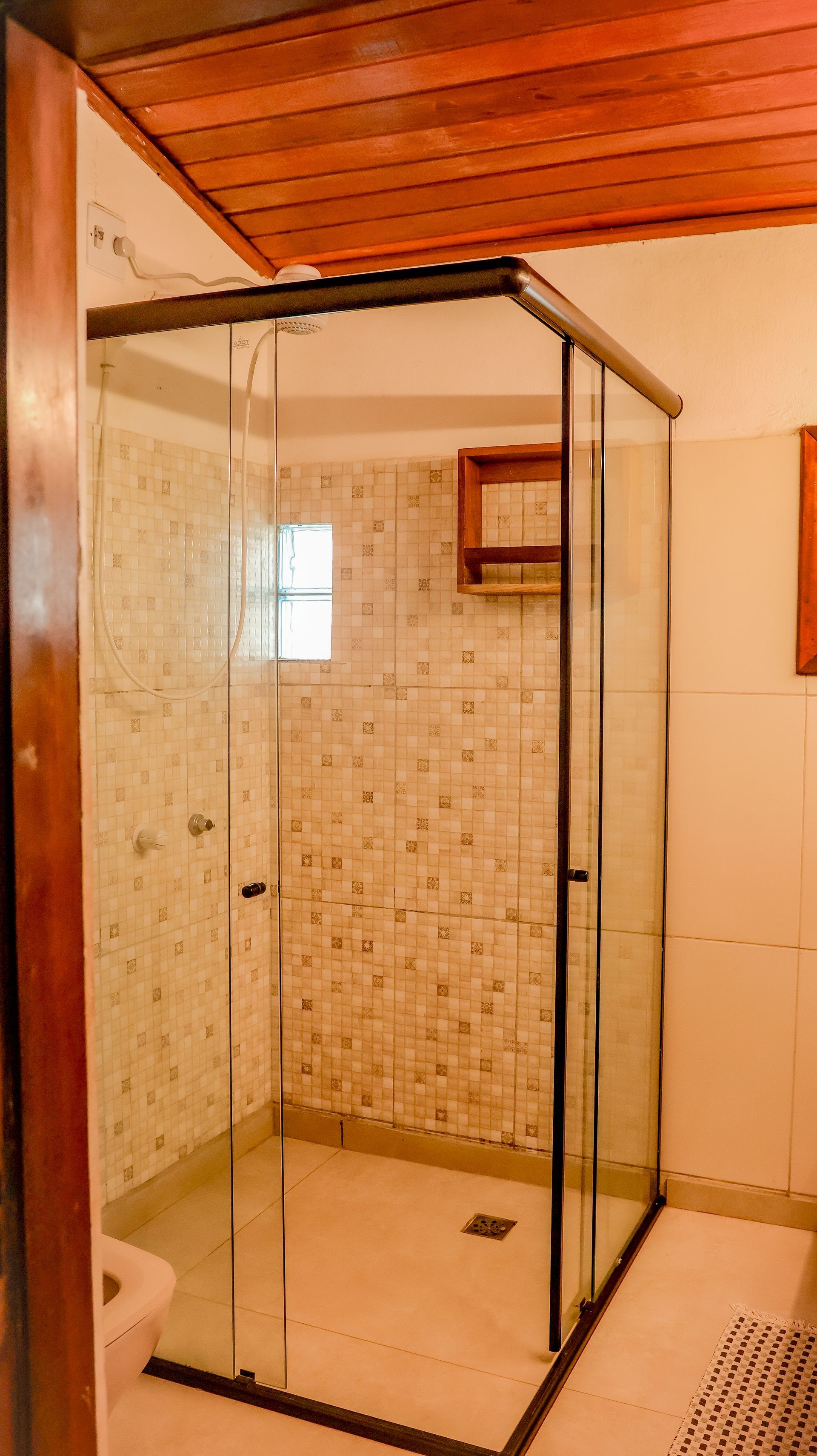 Box de vidro para chuveiro em um banheiro com paredes revestidas de azulejos em mosaico e teto de madeira.